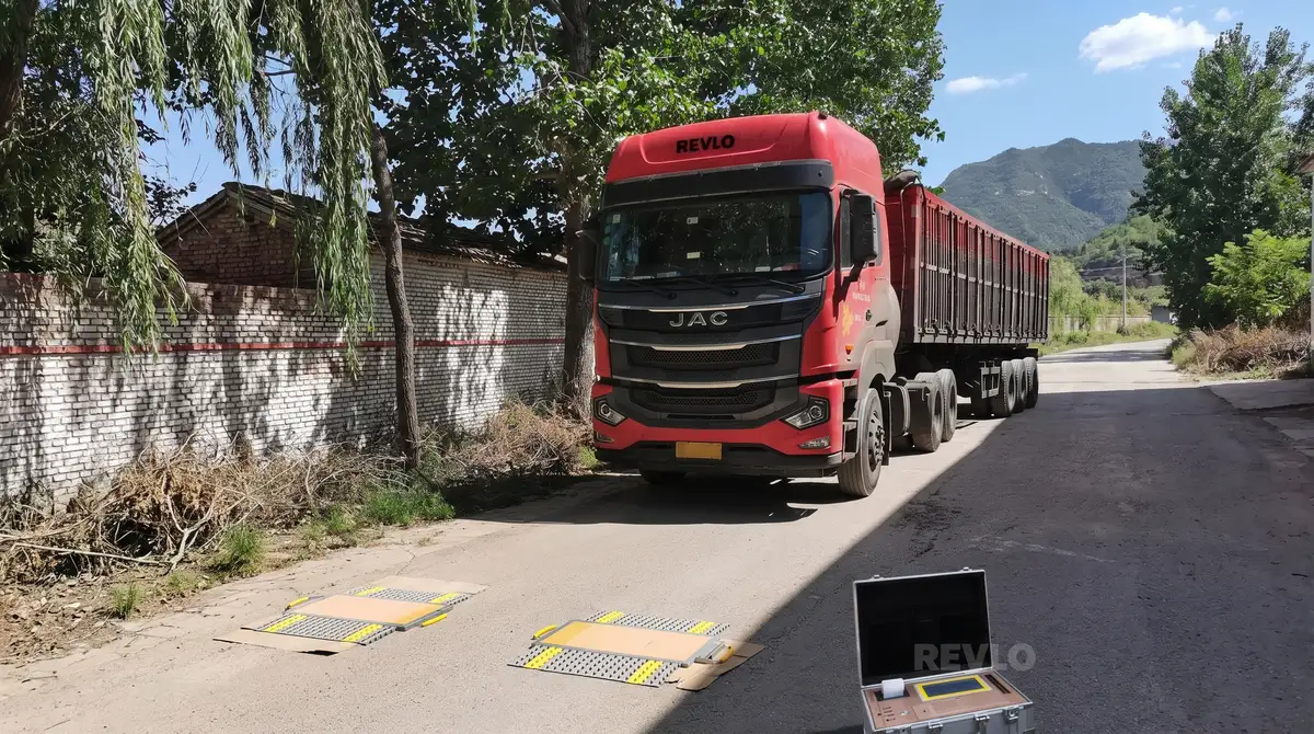 Caminhão com carreta basculante parado sobre as plataformas da balança rodoviária móvel, com a maleta do display aberta no chão ao lado
